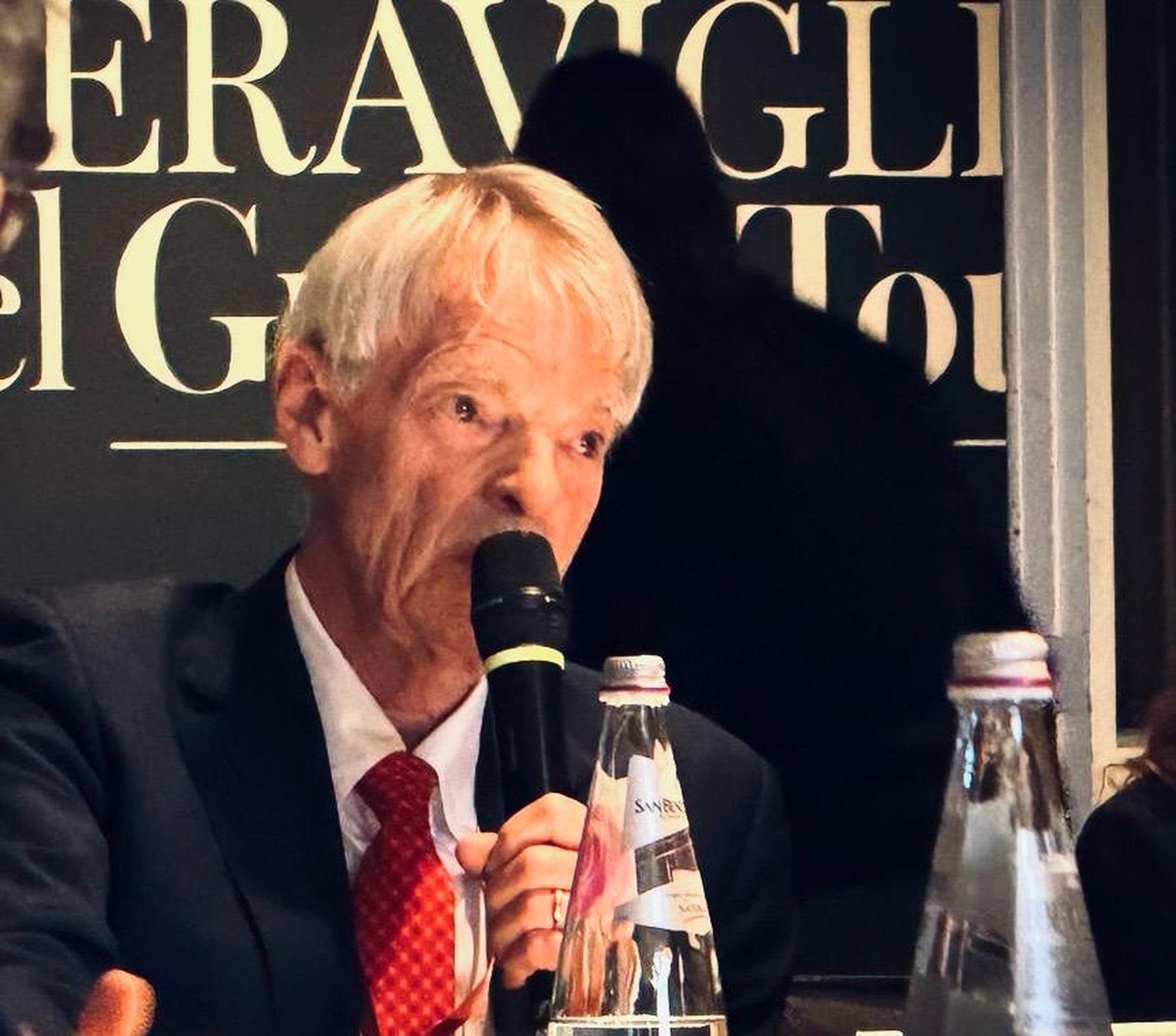 Michael Spence speaking into a microphone during The K.hub Journey roundtable on AI, growth and well-being, seated at a conference table with executives.